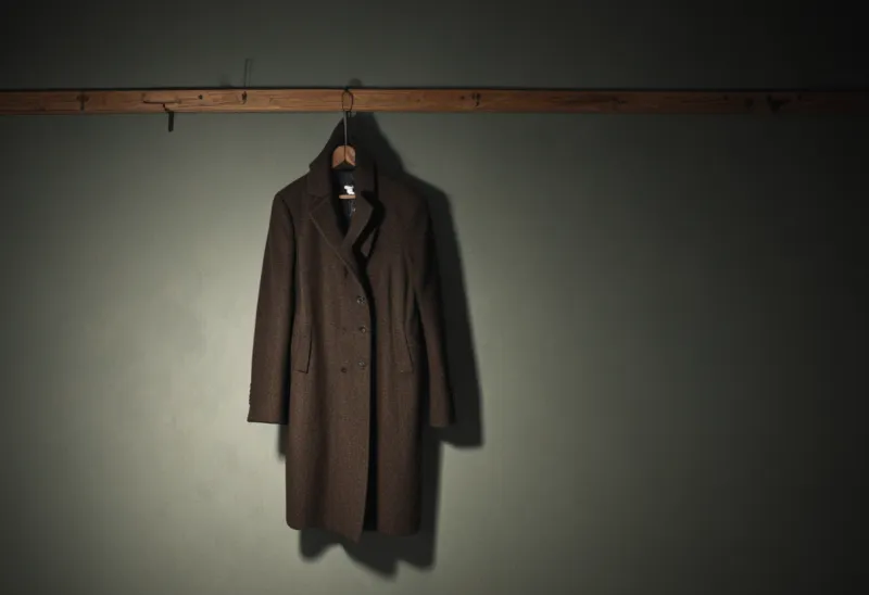 A single plain wool coat on a wooden hanger, hanging alone from a hook on a bare wall