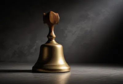 A polished brass hand bell with a wooden handle standing upright on a plain stone surface.