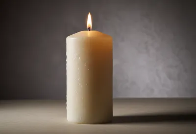 A white pillar candle with its small flame lit, on a stone surface.