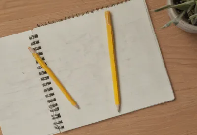 A closed spiral notebook with a yellow pencil resting on the cover, seen from above on a wooden desk